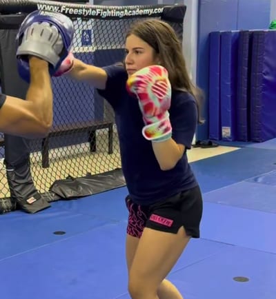 Camila De Las Cuevas in pink gloves throwing a punch into a coach's focus mitt inside the cage at Freestyle Fighting Academy