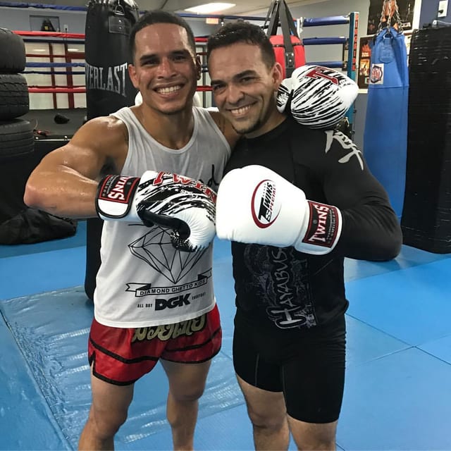 Chevenne Moreno photographed with a training partner in Muay Thai shorts and boxing gloves, arms around each other's shoulders beside the punching bags