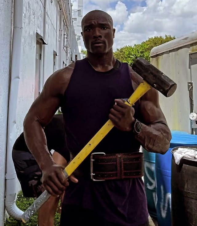 Emmanuel Lokolo outdoors in a lifting belt, holding a sledgehammer across his chest