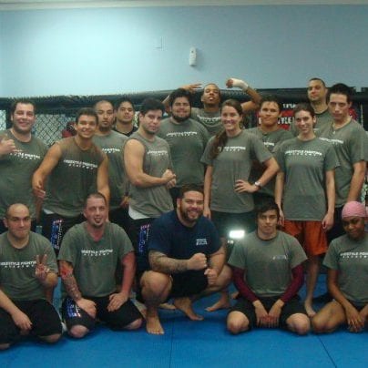 Un grupo grande de estudiantes adultos con camisetas de Freestyle Fighting Academy, de pie y arrodillados juntos en el tatami después de una clase de MMA en Miami