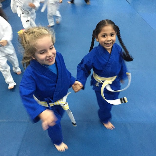 Dos niñas con kimonos azules de jiu-jitsu tomadas de la mano y corriendo sonrientes por el tatami en Freestyle Fighting Academy