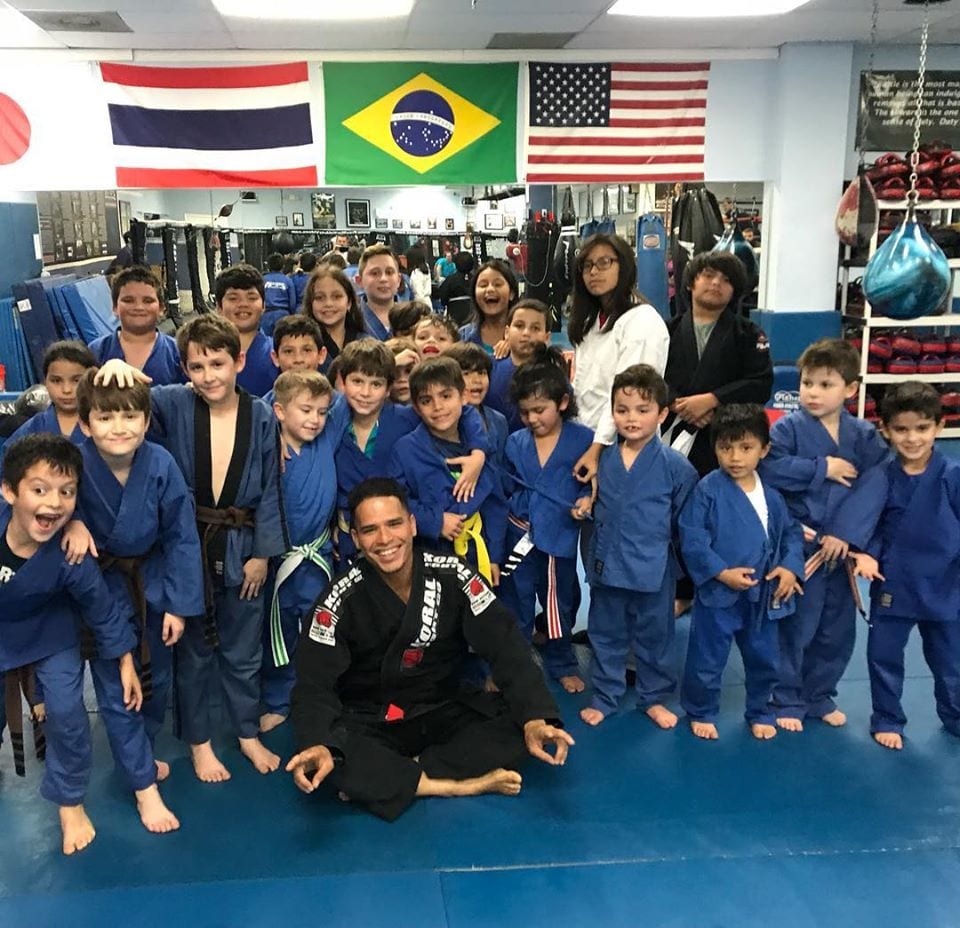Un grupo grande de niños con kimonos azules de jiu-jitsu posando con su entrenador en el tatami de Freestyle Fighting Academy, bajo las banderas de Tailandia, Brasil, Estados Unidos y Japón
