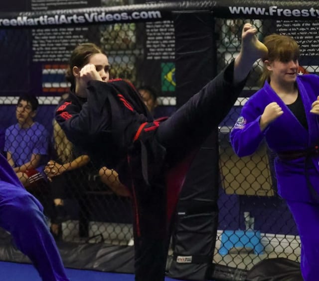 Krystal Cabrera in a black gi throwing a high kick inside the cage at Freestyle Fighting Academy