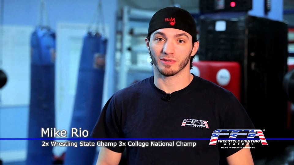 Mike Rio speaking to camera in front of the punching bags at Freestyle Fighting Academy, with an on-screen caption naming his wrestling titles